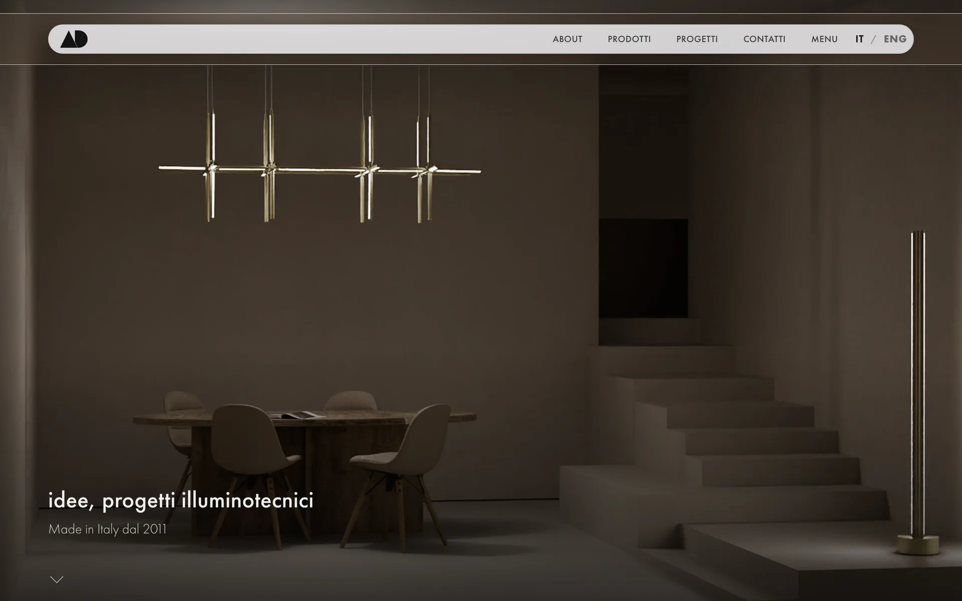 La home di adilluminazione.eu: lampada a sospensione su un tavolo di legno e scala in penombra, con il claim “idee, progetti illuminotecnici”